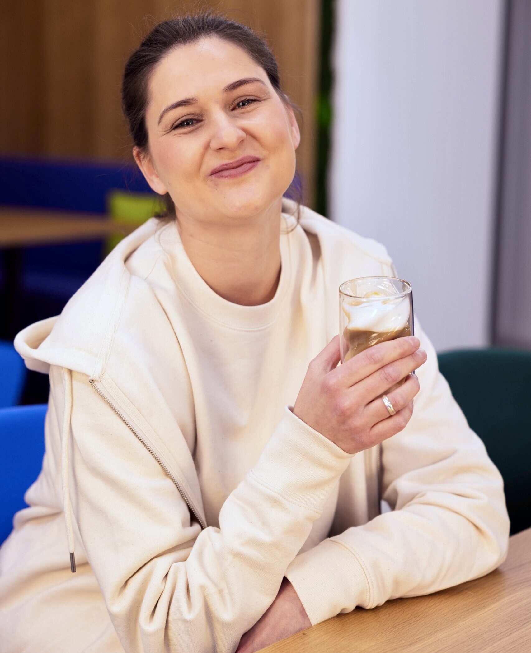 58BA6E3F-2E8F-4AF5-B5D8-7E1AB0EB9308_1_201_a Auf dem Bild sitzt eine Frau lächelnd an einem Tisch und trinkt einen Kaffee.